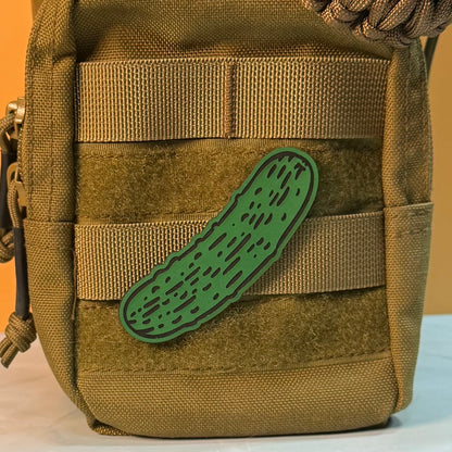 Dill Pickle laser-engraved synthetic morale patch: a green laser-engraved synthetic patch shaped like a cucumber or pickle with black textured lines, shown attached to the side webbing of a tan tactical bag.