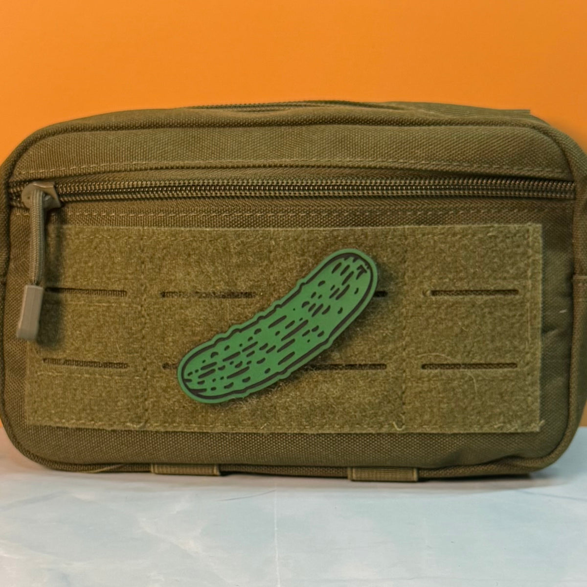 Dill Pickle laser-engraved synthetic morale patch: an olive green tactical pouch with a front hook-and-loop panel and a green cucumber-shaped patch, set against an orange background.
