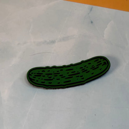 Dill Pickle laser-engraved synthetic morale patch: a dark green pickle-shaped patch or emblem with a black outline, resting on a light gray marble-patterned surface.