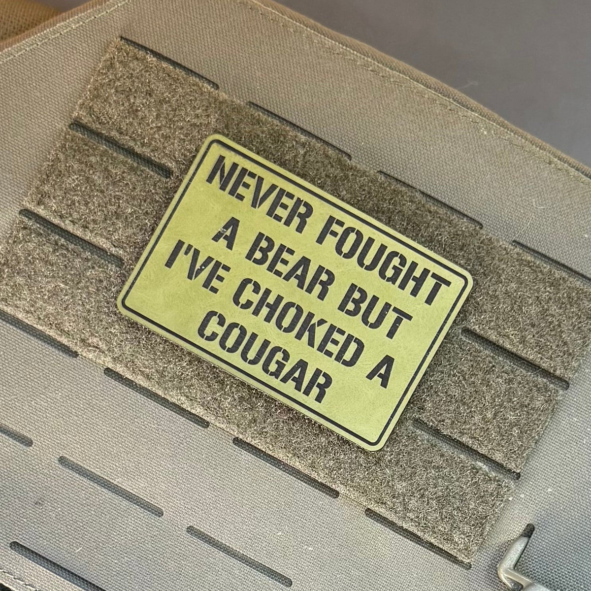 I’ve Choked A Cougar laser-engraved synthetic morale patch: a rectangular tactical patch with black text reading 'NEVER FOUGHT A BEAR BUT I'VE CHOKED A COUGAR' in olive green, shown attached to a tan MOLLE webbing strap on a.