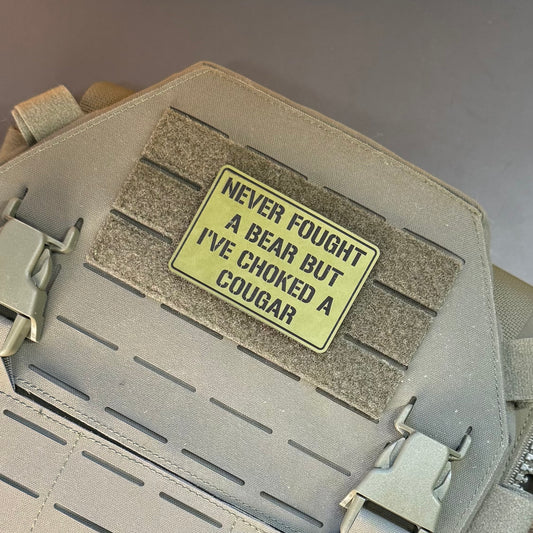 A rectangular tactical patch with black text reading 'NEVER FOUGHT A BEAR BUT I'VE CHOKED A COUGAR' in yellow-green, shown attached to a tan MOLLE webbing strap on a matching tactical bag.