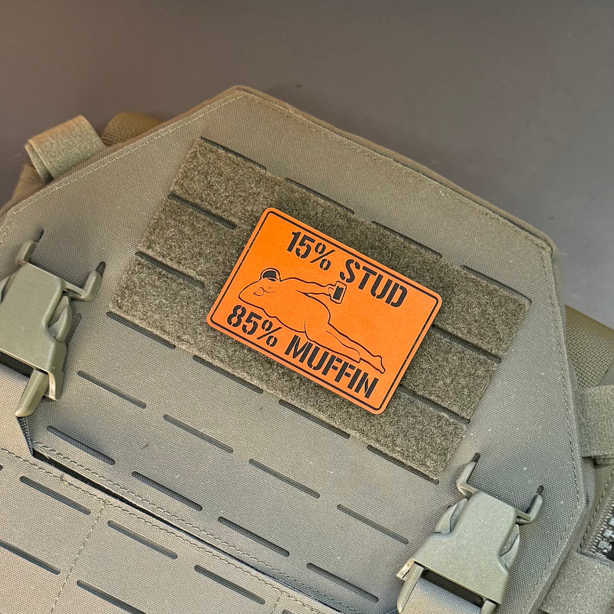 15% Stud 85% Muffin Funny Dad Bod laser-engraved synthetic morale patch: an orange rectangular patch with black text reading '15% STUD 85% MUFFIN' and a simple fish graphic, attached to a tan tactical vest or bag panel with velcro.