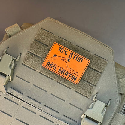 15% Stud 85% Muffin Funny Dad Bod laser-engraved synthetic morale patch: an orange rectangular patch with black text reading '15% STUD 85% MUFFIN' and a simple fish graphic, attached to a tan tactical vest or bag panel with velcro.