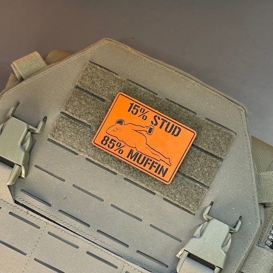 An orange rectangular patch with black text reading '15% STUD 85% MUFFIN' and a simple fish graphic, attached to a tan tactical vest or bag panel with velcro and buckles.