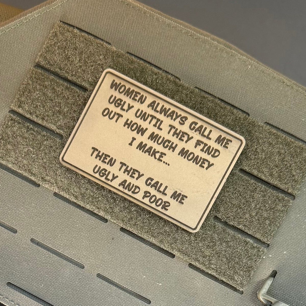 Women Call Me Ugly and Poor laser-engraved synthetic patch: a rectangular beige morale patch with black text, mounted on a gray hook-and-loop panel of a tactical vest. The patch reads: 'WOMEN ALWAYS GALL ME UGLY UNTIL THEY.