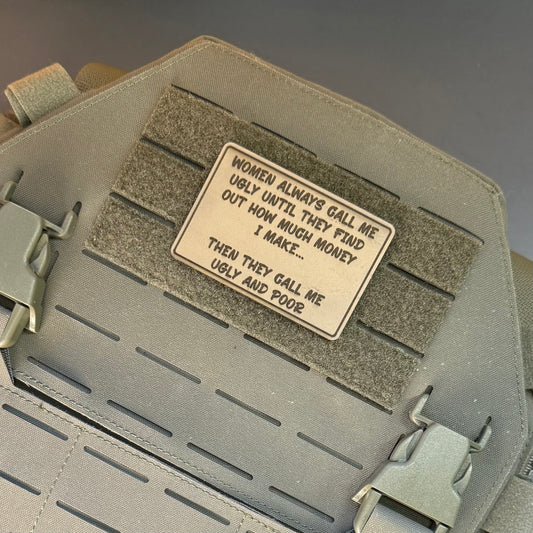 A rectangular beige morale patch with black text, mounted on a gray hook-and-loop panel of a tactical vest. The patch reads: 'WOMEN ALWAYS GALL ME UGLY UNTIL THEY FIND OUT HOW MUCH MONEY I MAKE.