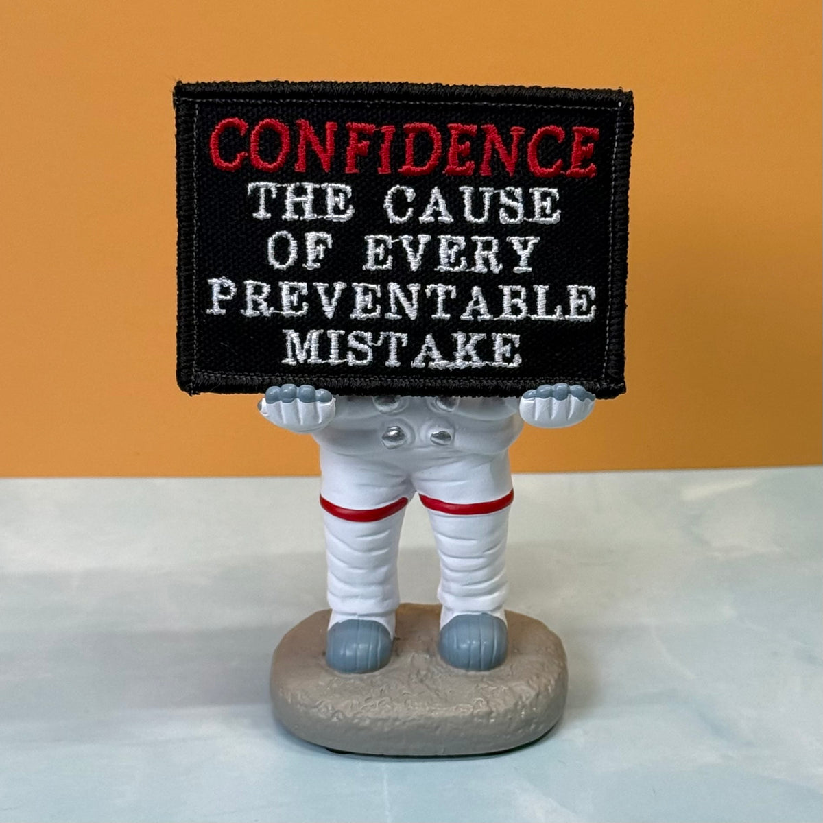 Confidence Causes Mistakes embroidered morale patch: small astronaut figurine holding a black embroidered sign that reads “CONFIDENCE THE CAUSE OF EVERY PREVENTABLE MISTAKE” in red and white lettering, set on a light surface.