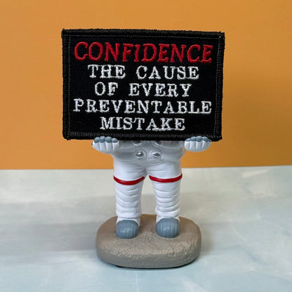 Confidence Causes Mistakes embroidered morale patch: small astronaut figurine holding a black embroidered sign that reads “CONFIDENCE THE CAUSE OF EVERY PREVENTABLE MISTAKE” in red and white lettering, set on a light surface.