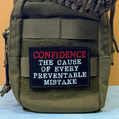 Confidence Causes Mistakes embroidered morale patch: olive green tactical pouch with a black embroidered patch that reads 'CONFIDENCE THE CAUSE OF EVERY PREVENTABLE MISTAKE' in red and white lettering.
