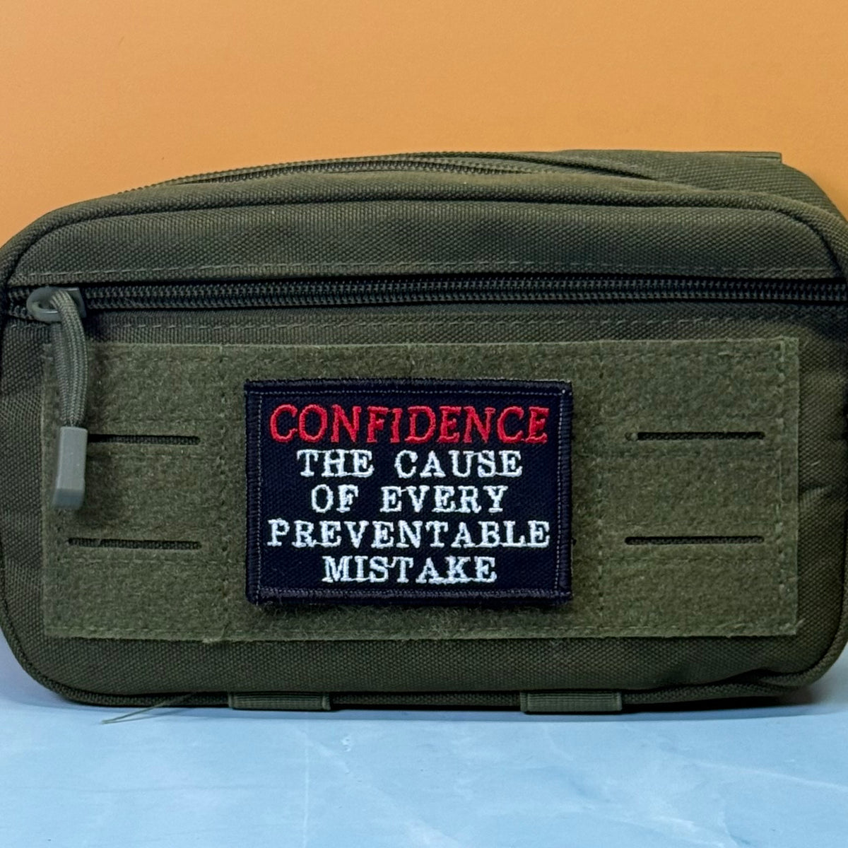 Confidence Causes Mistakes embroidered morale patch: olive green tactical pouch with a front hook-and-loop patch featuring the embroidered text 'CONFIDENCE THE CAUSE OF EVERY PREVENTABLE MISTAKE' in red and white on a black.