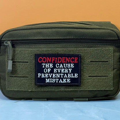 Confidence Causes Mistakes embroidered morale patch: olive green tactical pouch with a front hook-and-loop patch featuring the embroidered text 'CONFIDENCE THE CAUSE OF EVERY PREVENTABLE MISTAKE' in red and white on a black.