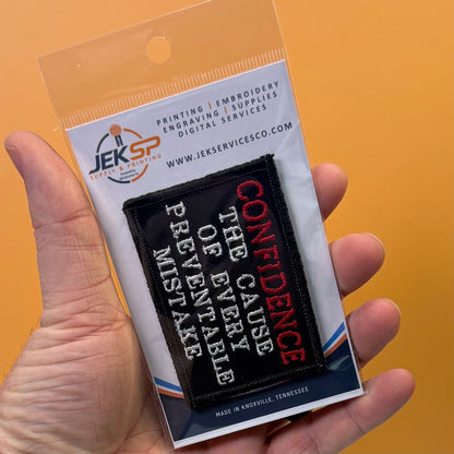 Confidence Causes Mistakes embroidered morale patch: hand holding a clear plastic blister pack containing a black embroidered patch with white and red text reading “CONFIDENCE THE CAUSE OF EVERY MISTAKE” on an orange background.