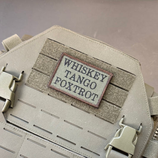 A close-up of a tactical vest or plate carrier in coyote tan, featuring a rectangular embroidered patch that reads 'WHISKEY TANGO FOXTROT' in dark gray letters with a red border, mounted on a Velcro patch panel.