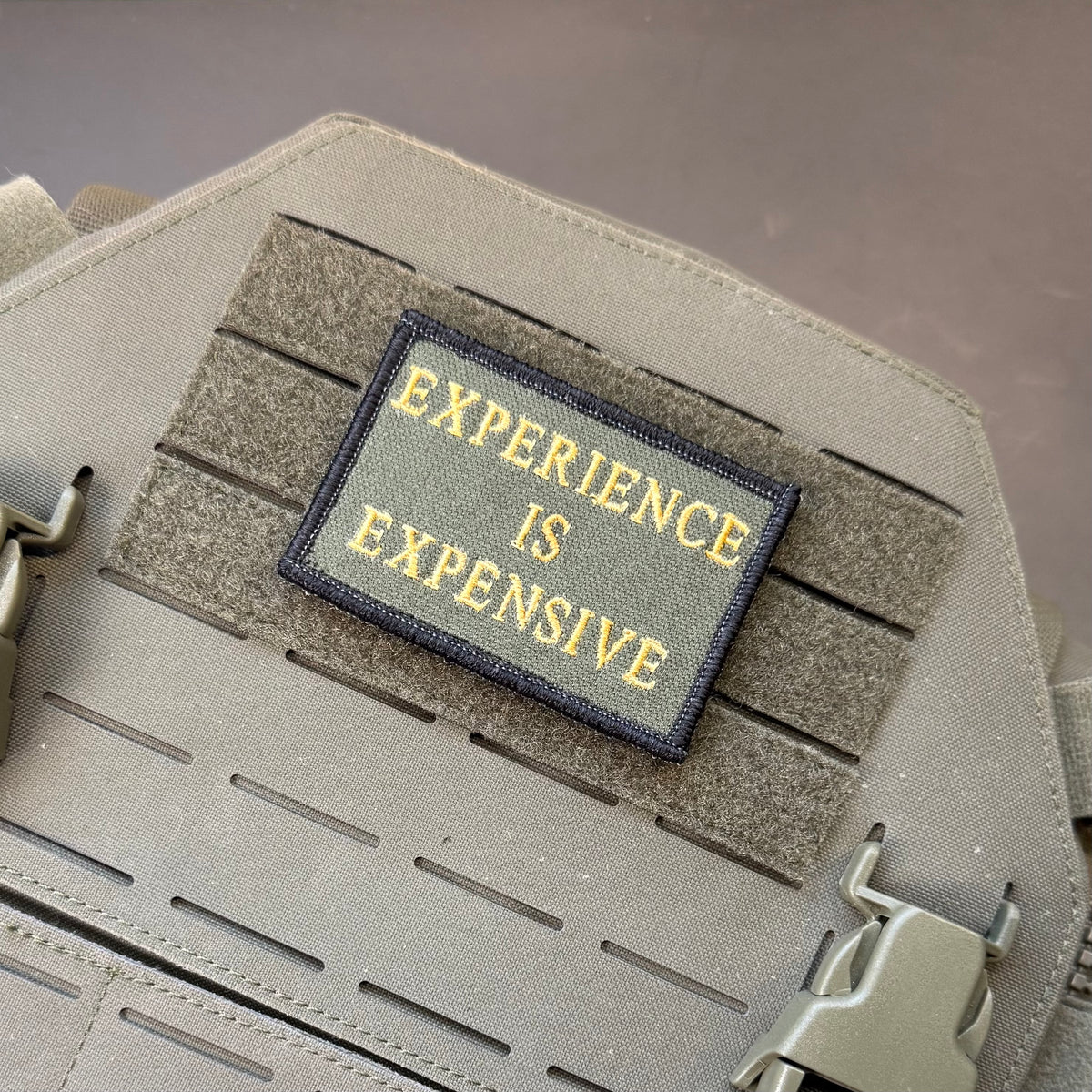 Experience Is Expensive embroidered morale patch: with the words 'EXPERIENCE IS EXPENSIVE' in yellow lettering on a dark olive background, attached to a gray tactical vest with MOLLE webbing.