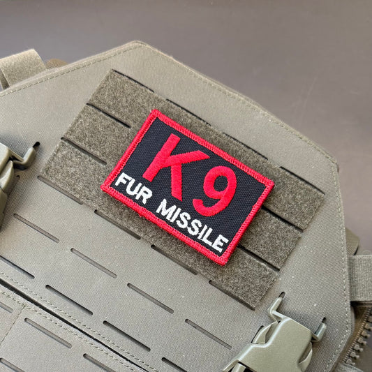 A rectangular tactical patch with red and white embroidery reading 'K9 FUR MISSILE' on a black background, attached to a tan MOLLE tactical vest.
