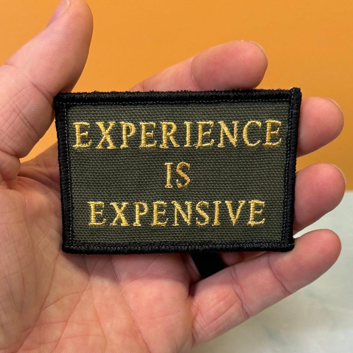 Experience Is Expensive embroidered morale patch: held in a hand, featuring the text 'EXPERIENCE IS EXPENSIVE' in gold thread on an olive green background with a black stitched border.