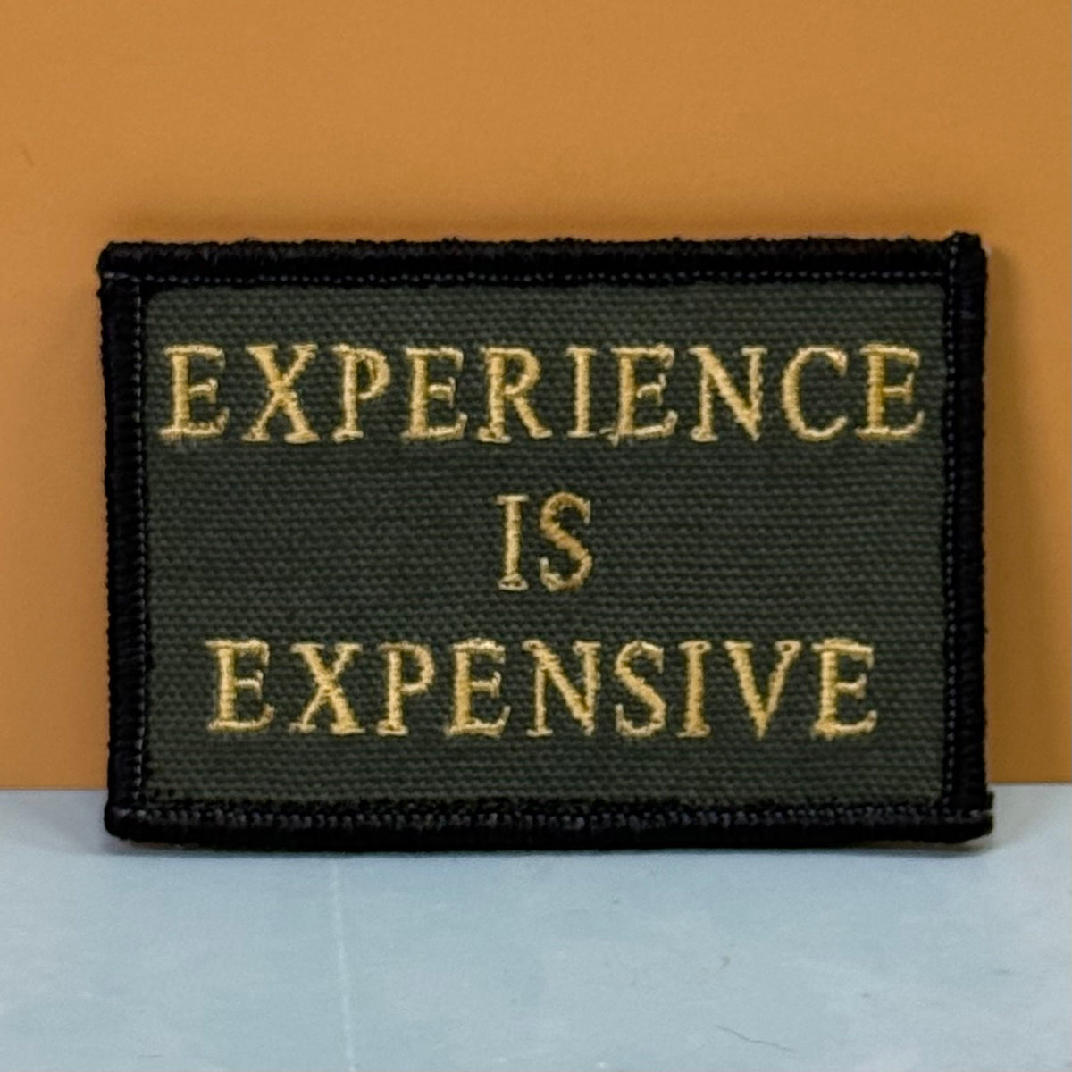 Experience Is Expensive embroidered morale patch: rectangular patch with the phrase 'EXPERIENCE IS EXPENSIVE' in gold lettering on a dark olive green background, finished with a black stitched border, set against a warm.