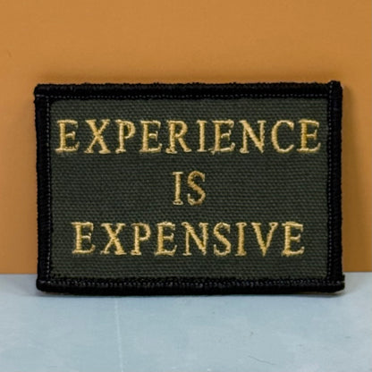 Experience Is Expensive embroidered morale patch: rectangular patch with the phrase 'EXPERIENCE IS EXPENSIVE' in gold lettering on a dark olive green background, finished with a black stitched border, set against a warm.