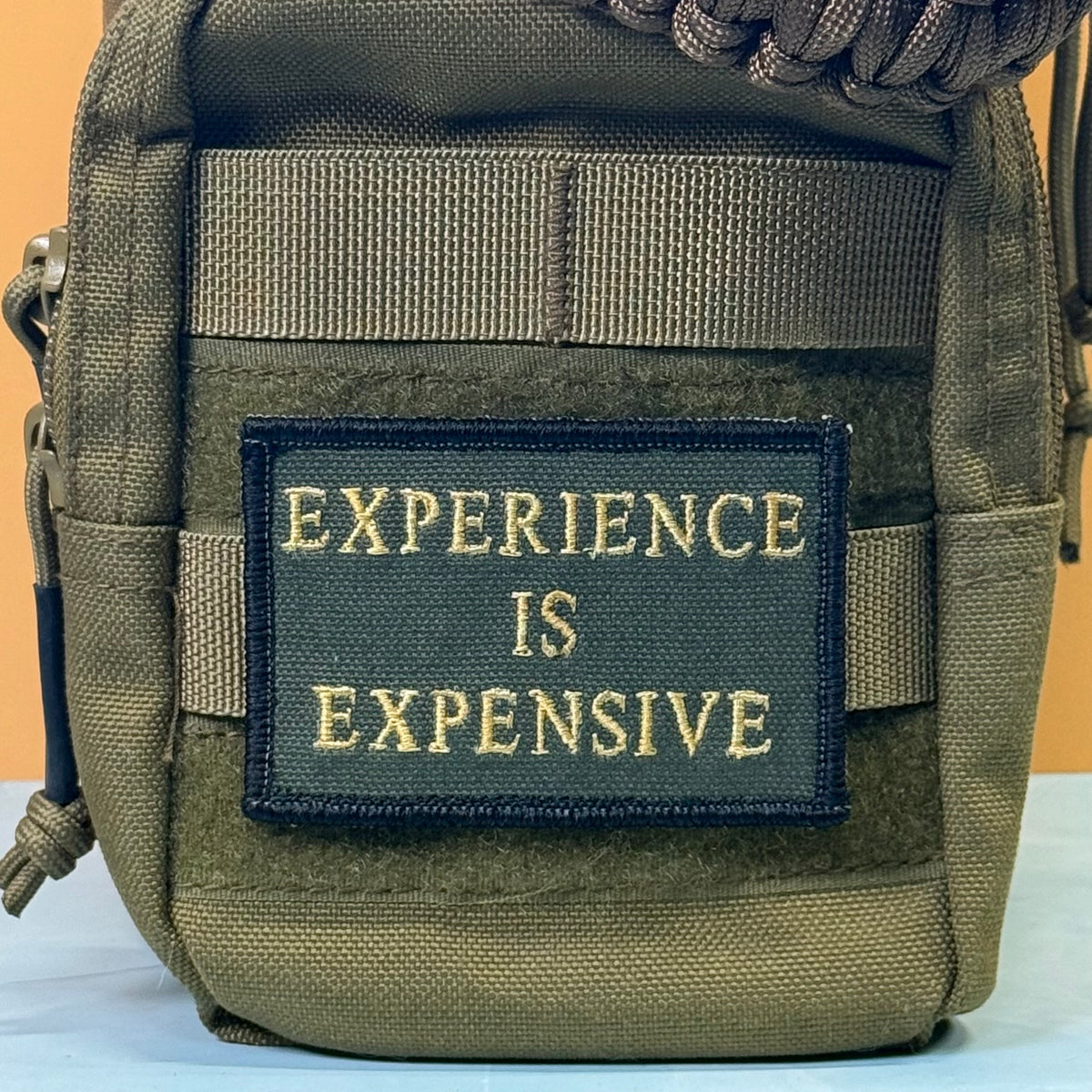 Experience Is Expensive embroidered morale patch: olive green tactical pouch with a black rectangular patch featuring the gold embroidered text 'EXPERIENCE IS EXPENSIVE'.
