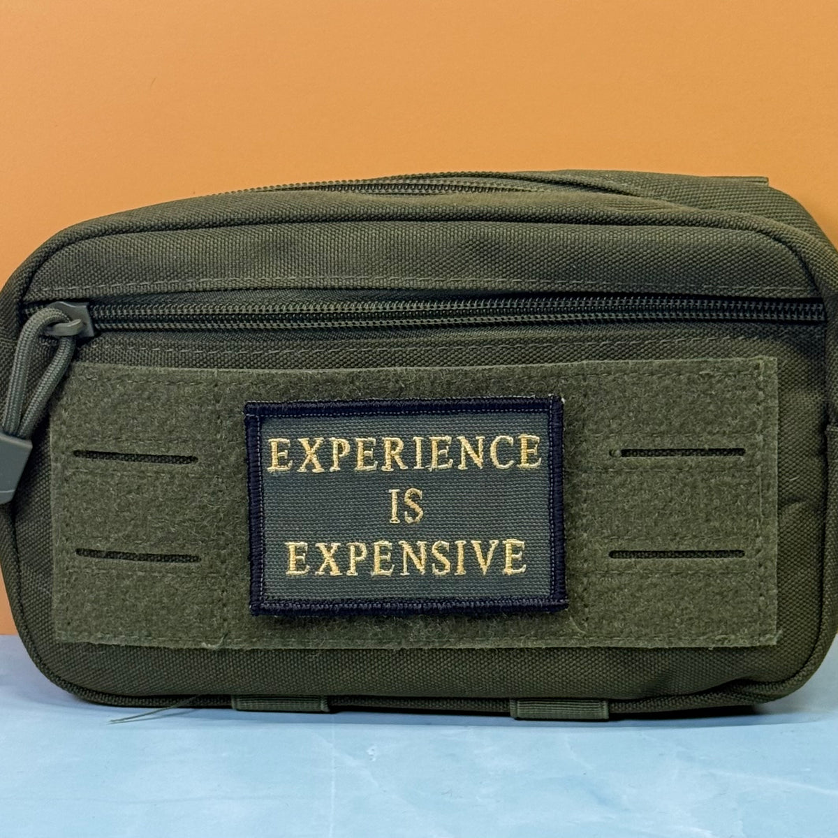 Experience Is Expensive embroidered morale patch: olive green tactical pouch with a front patch reading 'EXPERIENCE IS EXPENSIVE' in gold lettering, set against a split orange and light blue background.