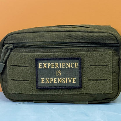 Experience Is Expensive embroidered morale patch: olive green tactical pouch with a front patch reading 'EXPERIENCE IS EXPENSIVE' in gold lettering, set against a split orange and light blue background.