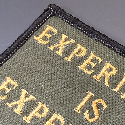 Experience Is Expensive embroidered morale patch: showing olive green fabric with black stitched border and gold/yellow text reading 'EXPERI.