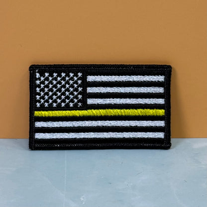Thin Yellow Line American Flag embroidered morale patch: American flag patch with a thin yellow line, featuring black, white, and yellow embroidery on a black background, displayed against a split orange and gray background.