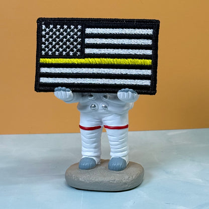 Thin Yellow Line American Flag embroidered morale patch: astronaut figurine holding a black and white American flag patch with a single yellow stripe, set against a two-toned background of orange and light gray.