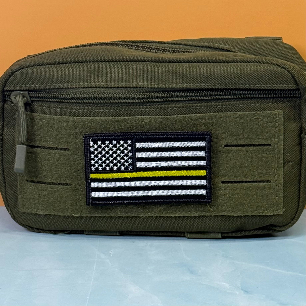 Thin Yellow Line American Flag embroidered morale patch: olive drab tactical pouch with a front hook-and-loop patch featuring a black and white American flag with a single yellow stripe, set against an orange and light blue background.