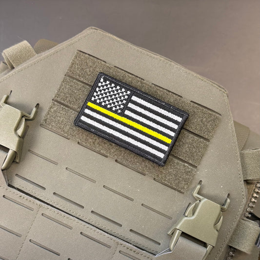 A tactical patch featuring a black and white American flag with a single yellow stripe, mounted on a tan MOLLE tactical vest.
