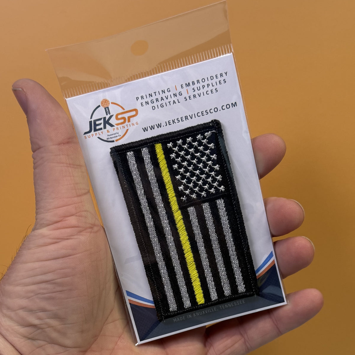 Thin Yellow Line American Flag embroidered morale patch: hand holding a packaged embroidered patch featuring a black American flag with a single yellow stripe, on a white backing card from Jeksp Supplies Co. against a solid orange background.