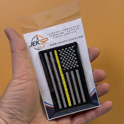 Thin Yellow Line American Flag embroidered morale patch: hand holding a packaged embroidered patch featuring a black American flag with a single yellow stripe, on a white backing card from Jeksp Supplies Co. against a solid orange background.