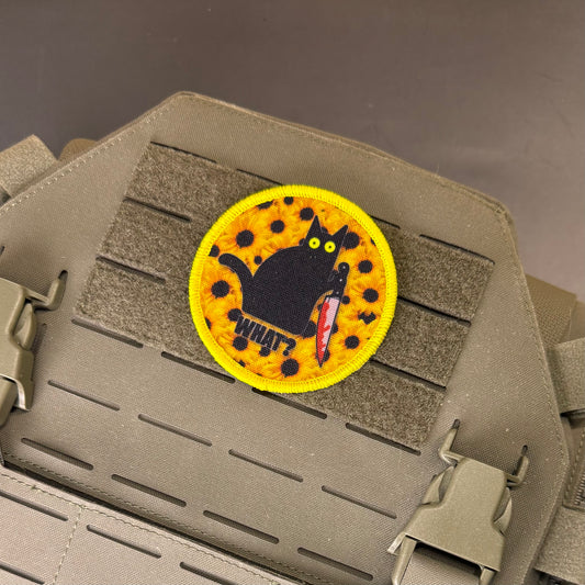 A circular embroidered tactical patch featuring a black cat holding a red knife on a yellow spotted background with the word 'WHY?' below it, attached to a tan tactical vest with Velcro and quick-release buckles.