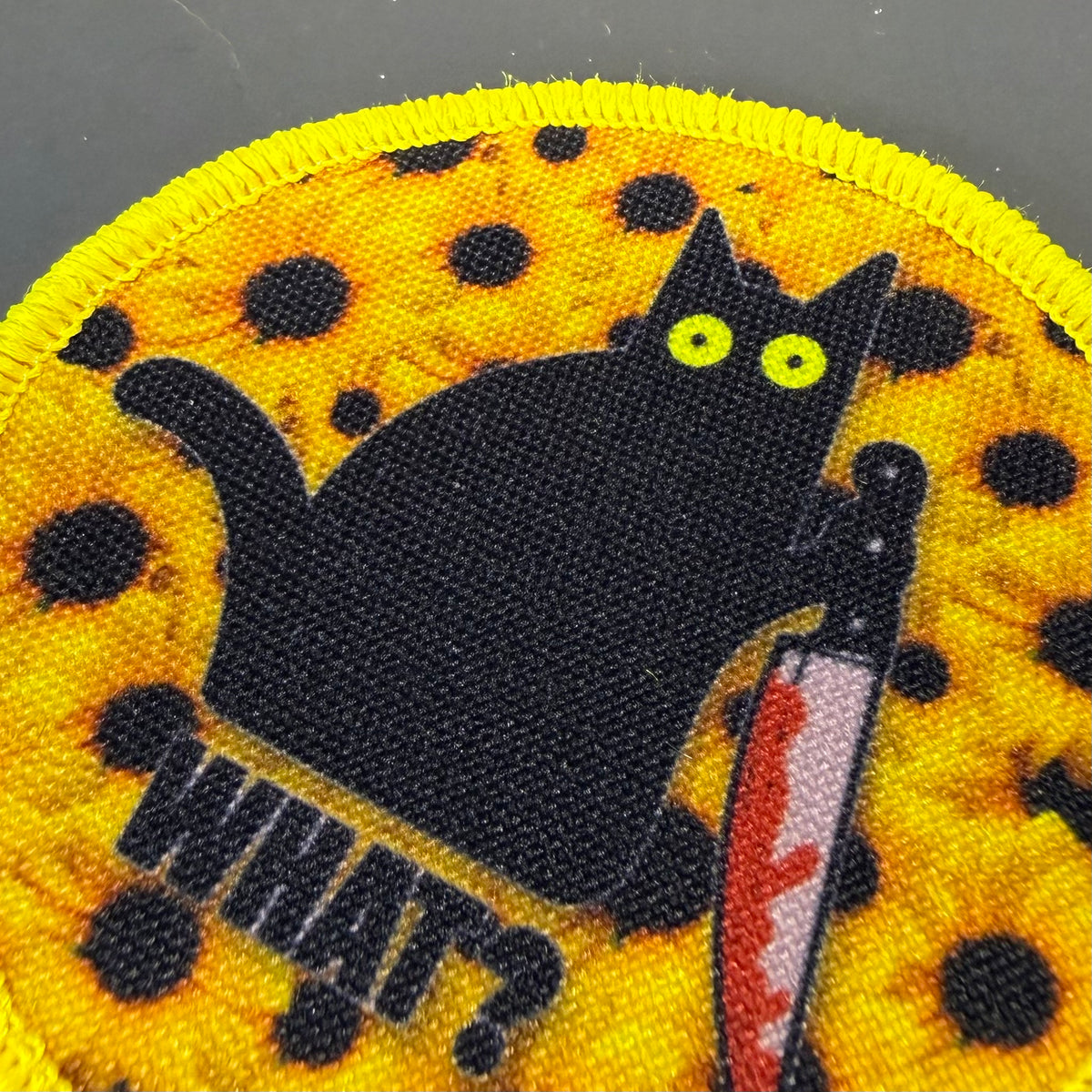Murder Cat and Sunflowers printed canvas morale patch, a close-up of a circular printed patch featuring a black cat holding a bloody knife, with the design lettering in black text on a yellow background with black spots.