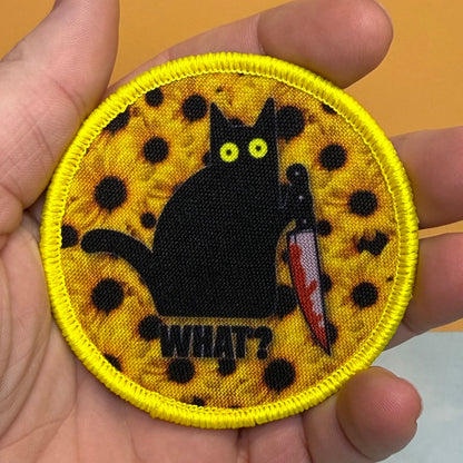 Murder Cat and Sunflowers printed canvas morale patch held in a hand, featuring a black cat holding a bloody knife on a yellow spotted background with the design lettering below it.