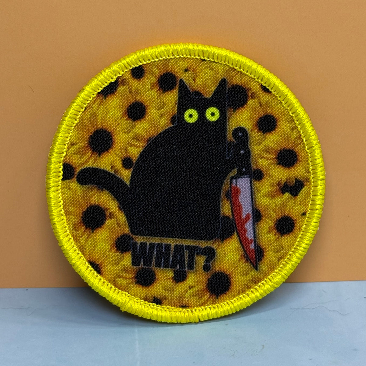 Murder Cat and Sunflowers printed canvas morale patch featuring a black cat holding a bloody knife, with the design lettering below it. The patch has a bright yellow border and a yellow background with black spots, set against.