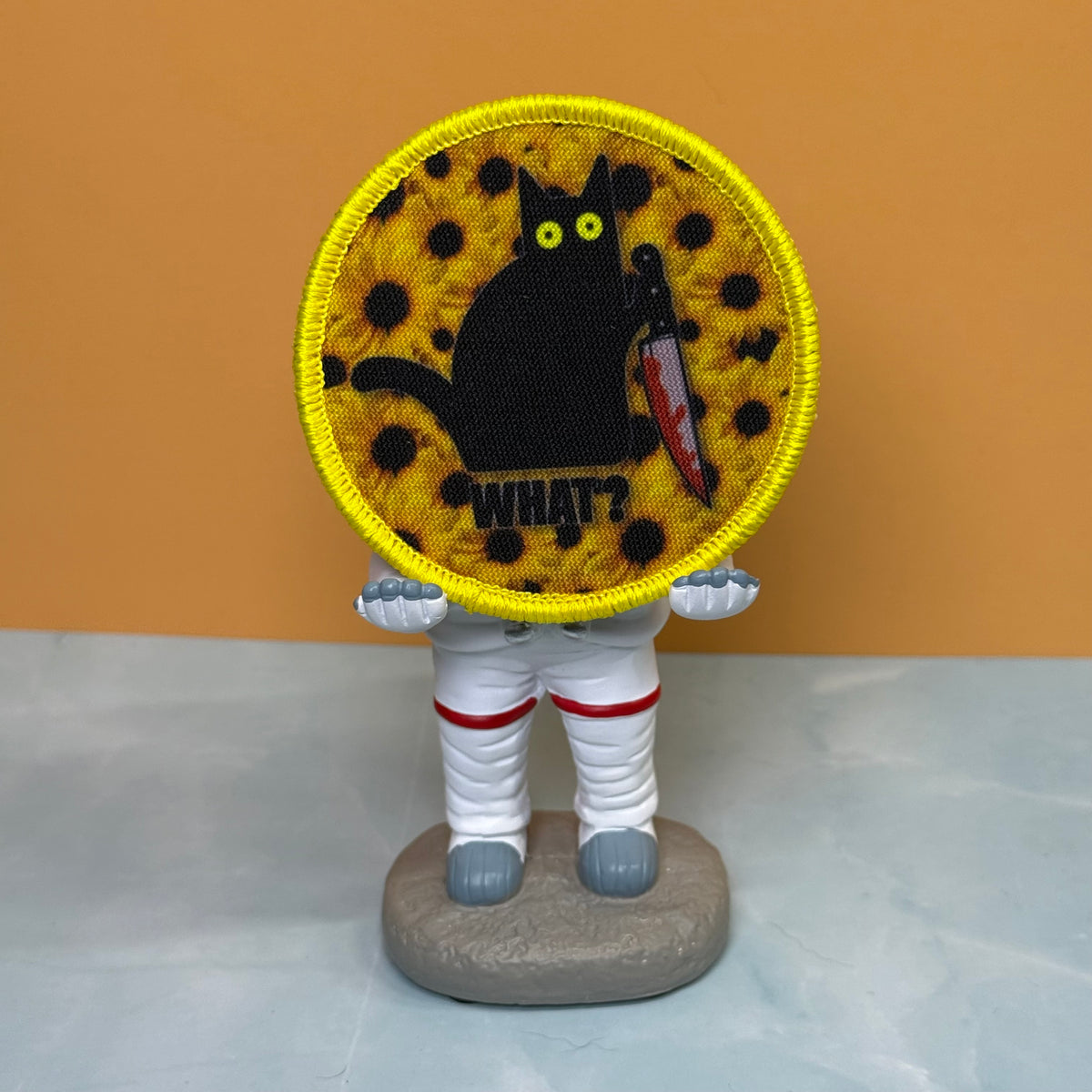 Murder Cat and Sunflowers printed canvas morale patch, a small astronaut figurine standing on a gray base, holding a round printed patch in front of its face. The patch shows a black cat on a yellow patterned background with.