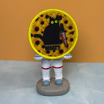Murder Cat and Sunflowers printed canvas morale patch, a small astronaut figurine standing on a gray base, holding a round printed patch in front of its face. The patch shows a black cat on a yellow patterned background with.