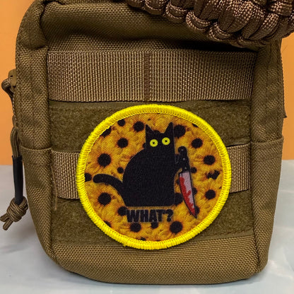 Murder Cat and Sunflowers printed canvas morale patch, a close-up of a tactical pouch in olive drab brown nylon, featuring a circular printed patch with a black cat holding a bloody knife on a yellow spotted background with.