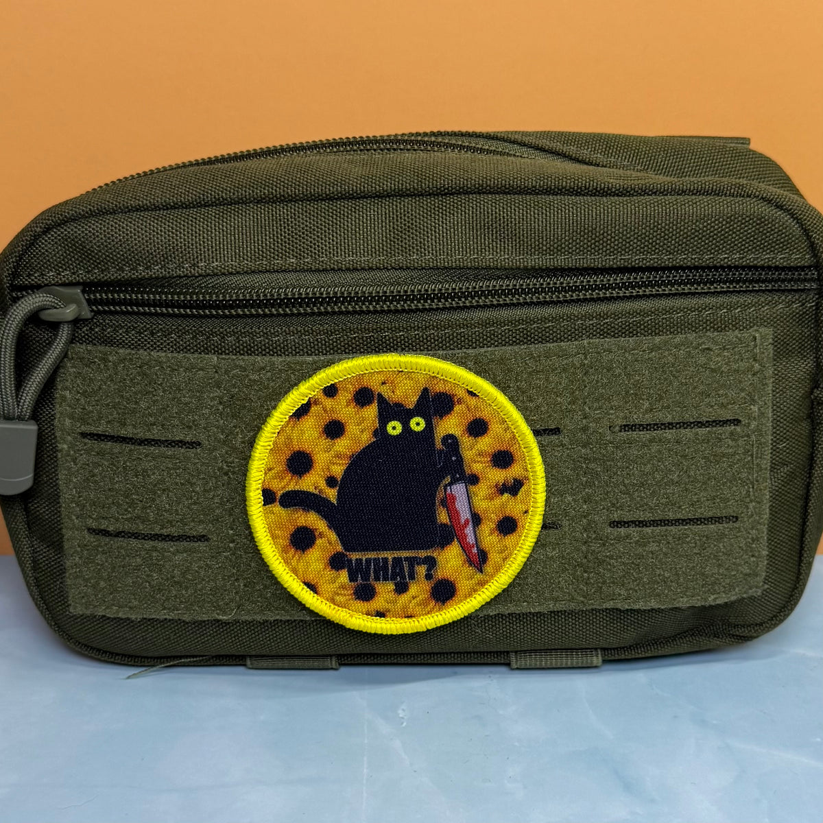 Murder Cat and Sunflowers printed canvas morale patch, an olive green tactical pouch with a hook-and-loop front panel, featuring a circular printed patch of a black cat holding a knife on a yellow spotted background with the.