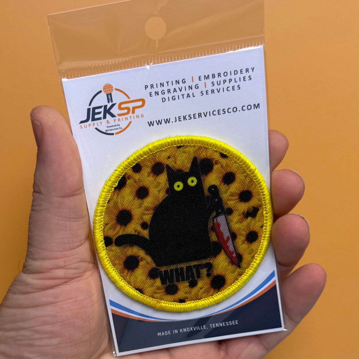 Murder Cat and Sunflowers printed canvas morale patch, a hand holds a packaged printed patch featuring a black cat on a yellow spotted background with the design lettering below it. The patch is mounted on a white backing card.