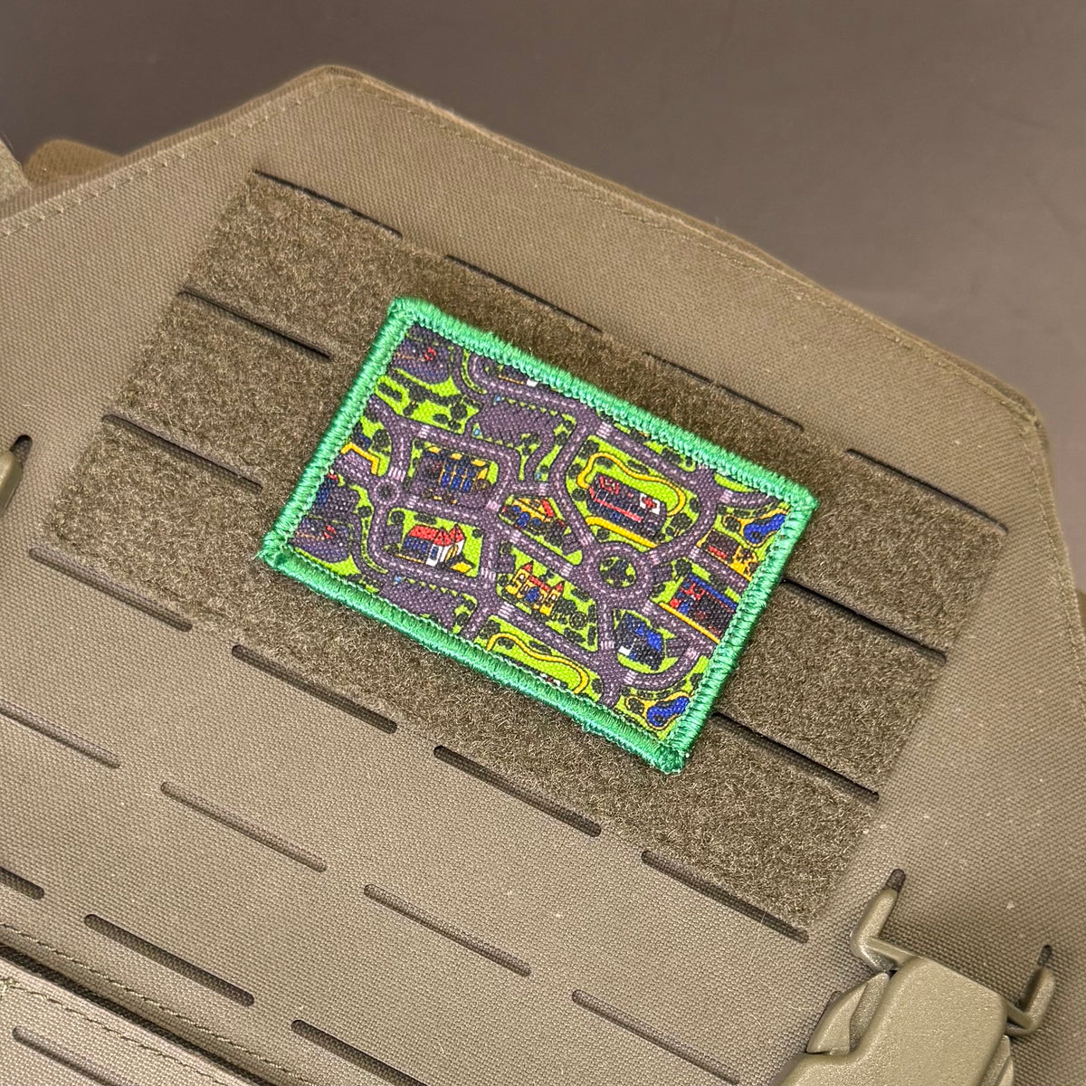 A rectangular tactical patch featuring a colorful street map design with a bright green border, shown attached to a tan tactical vest with Velcro loops.