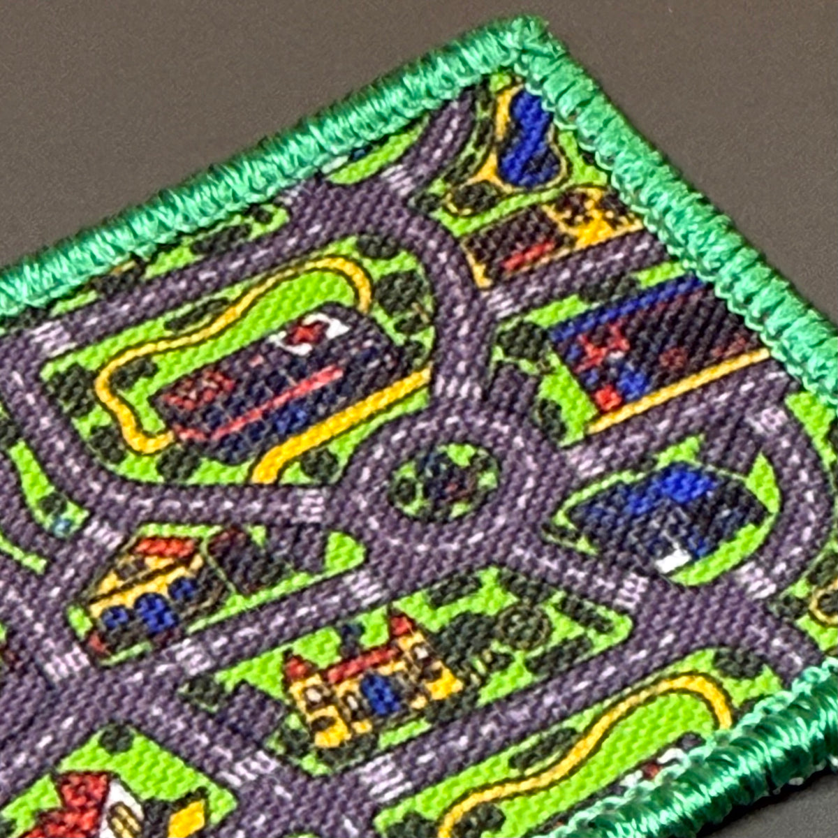 A close-up of a square embroidered patch featuring a colorful road and traffic pattern with cars, houses, and green grass, finished with a green stitched border.