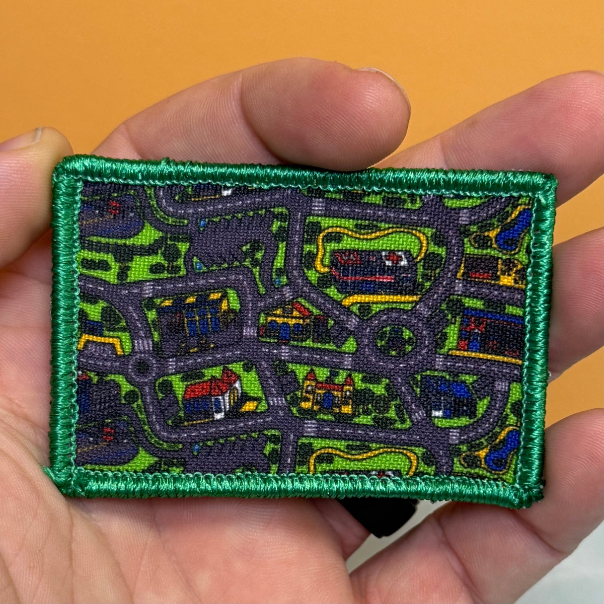 A rectangular embroidered patch with a green border and a detailed street-map pattern in green, gray, blue, red, and yellow, held in a person's hand against a solid orange background.