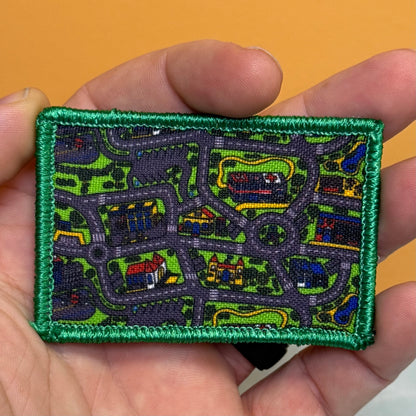 A rectangular embroidered patch with a green border and a detailed street-map pattern in green, gray, blue, red, and yellow, held in a person's hand against a solid orange background.