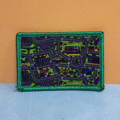 A rectangular embroidered patch featuring a street map design with houses, roads, and green areas, finished with a thick green border, shown against a two-tone orange and light blue background.