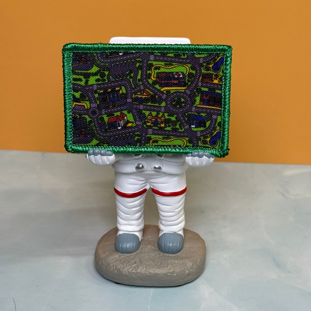 A novelty astronaut figurine holding a large embroidered patch with a street-map design and green border, standing on a gray base against an orange and gray background.