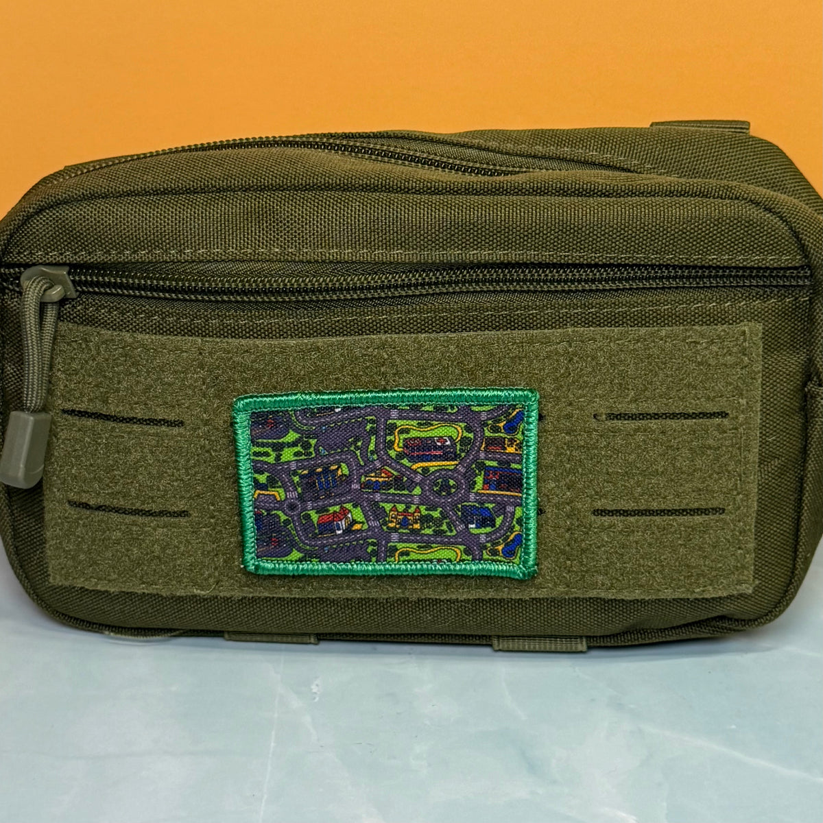 An olive green tactical pouch with a top zipper and a rectangular embroidered patch featuring a multicolor map design with a green border, set against an orange background.