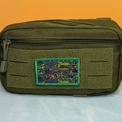 An olive green tactical pouch with a top zipper and a rectangular embroidered patch featuring a multicolor map design with a green border, set against an orange background.
