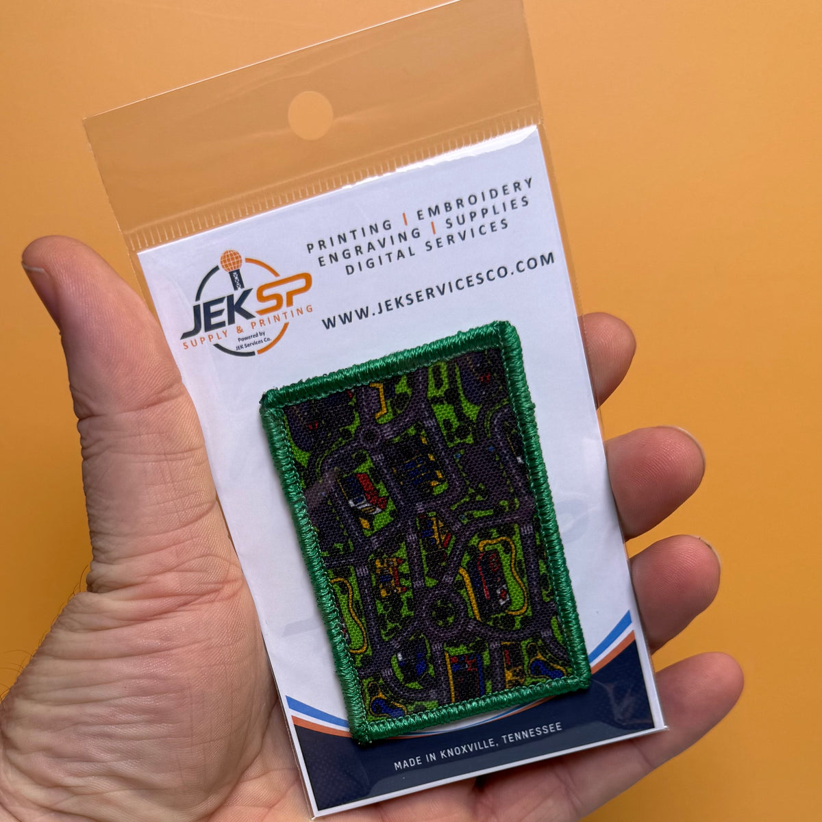A hand holds a clear plastic bag containing a small embroidered patch on a branded white backing card. The patch has a green border and a dark abstract map-like design with bright green, yellow, red, and blue details.